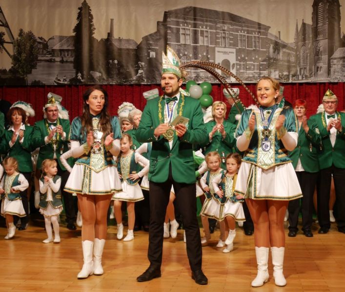 Eine Tanzgarde mit Kindern und Erwachsenen auf der Bühne, mittig steht ein Mann mit Narrenkappe und Mikrofon in der Hand.