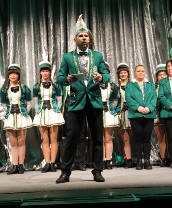 Ein Mann in grünem Sakko spricht auf Bühne, flankiert von Gardetänzerinnen und Team in Uniform.