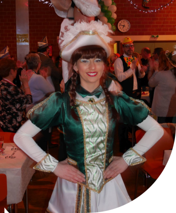 Eine junge Frau in grün-weißer Gardetracht steht lächelnd in einem Festsaal mit Publikum und Karnevalsdekoration.