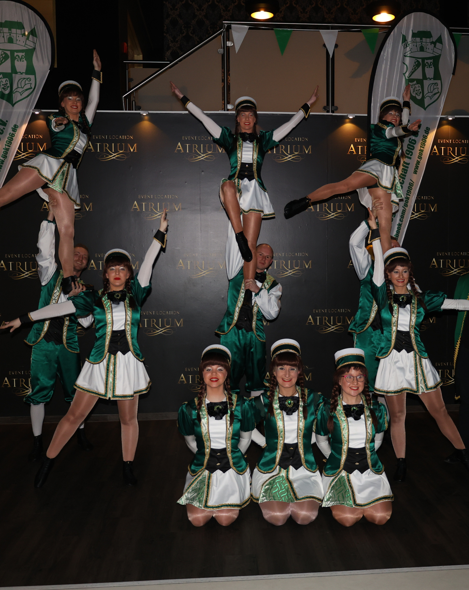 Gardegruppe zeigt akrobatische Hebefiguren in grünen Kostümen vor einem schwarzen Bühnenhintergrund mit Vereinslogo.