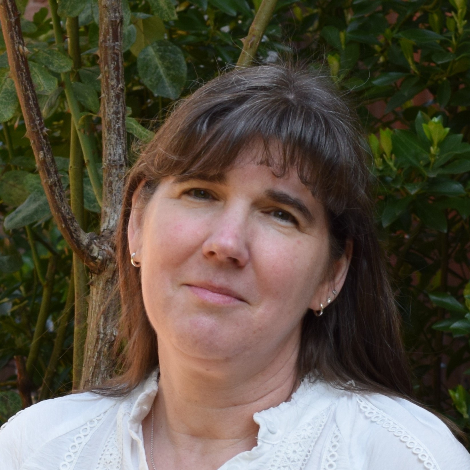 Frau mit langen braunen Haaren blickt ruhig in weißer Bluse vor einem grünen Busch in die Kamera.