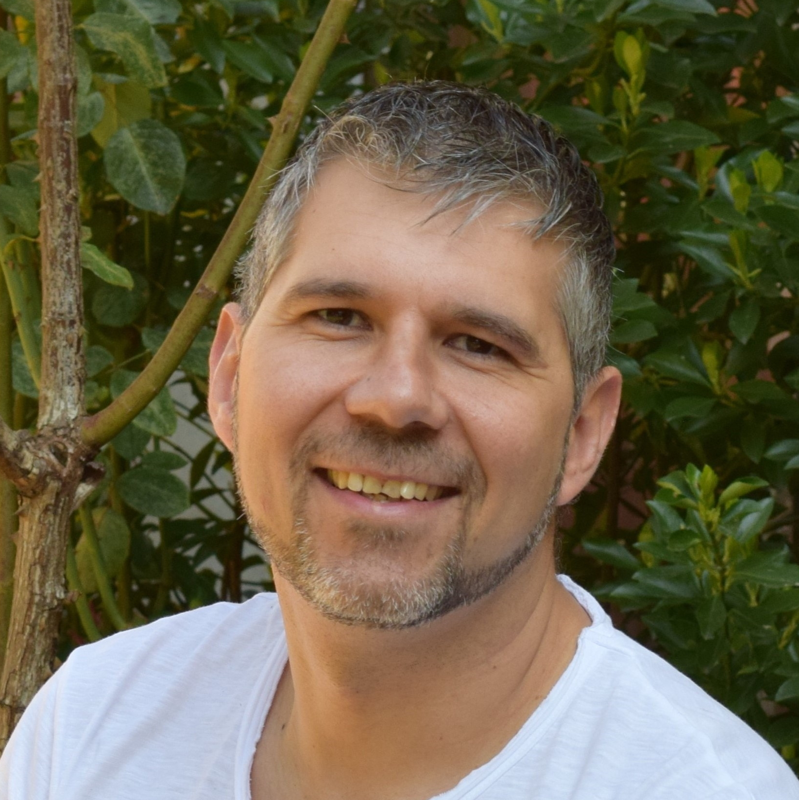 Mann mit Bart und weißem Hemd lächelt freundlich vor grünem Blätterhintergrund in die Kamera.