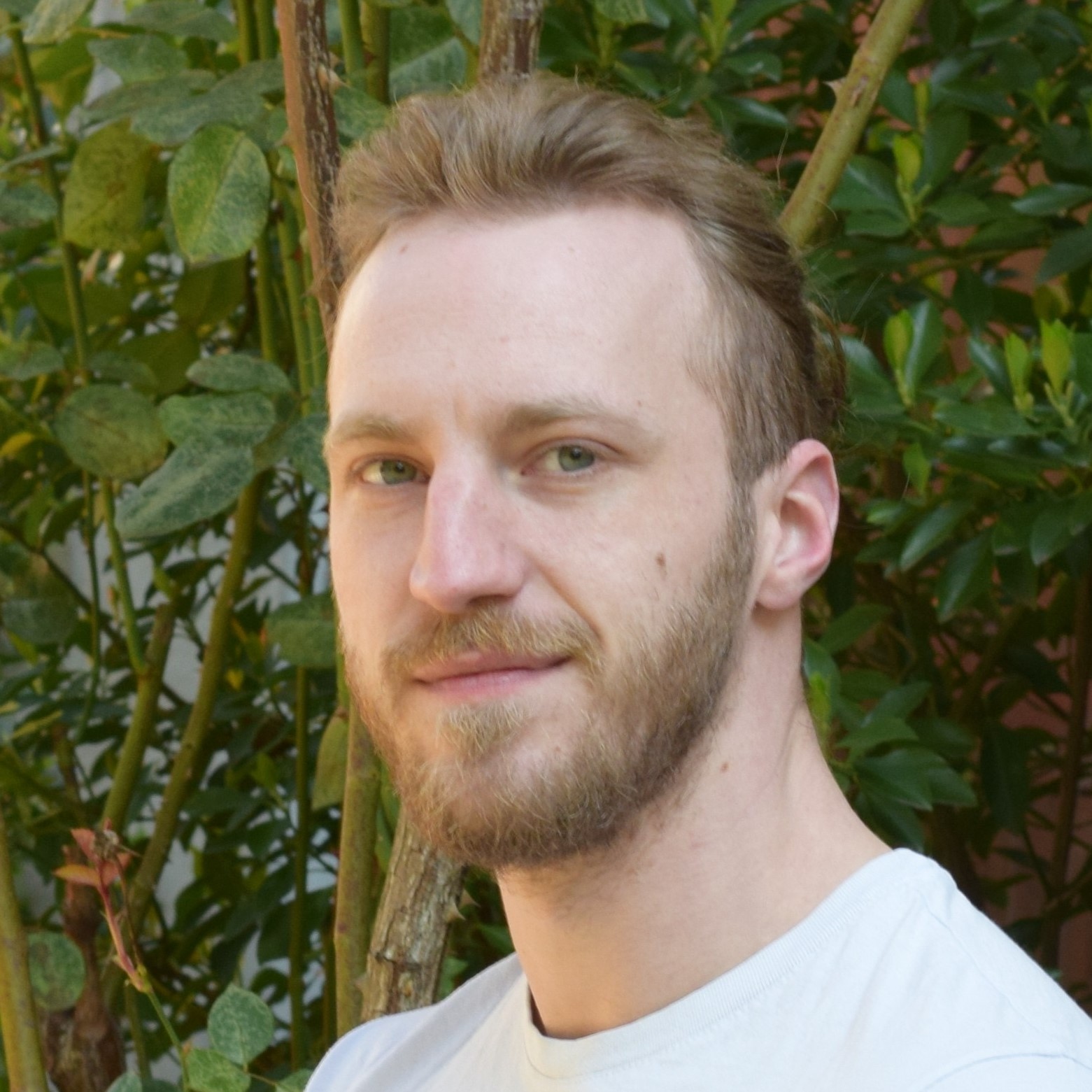 Junger Mann mit zurückgekämmtem Haar, Bart und weißem Shirt blickt ruhig in die Kamera, grüner Busch im Hintergrund.