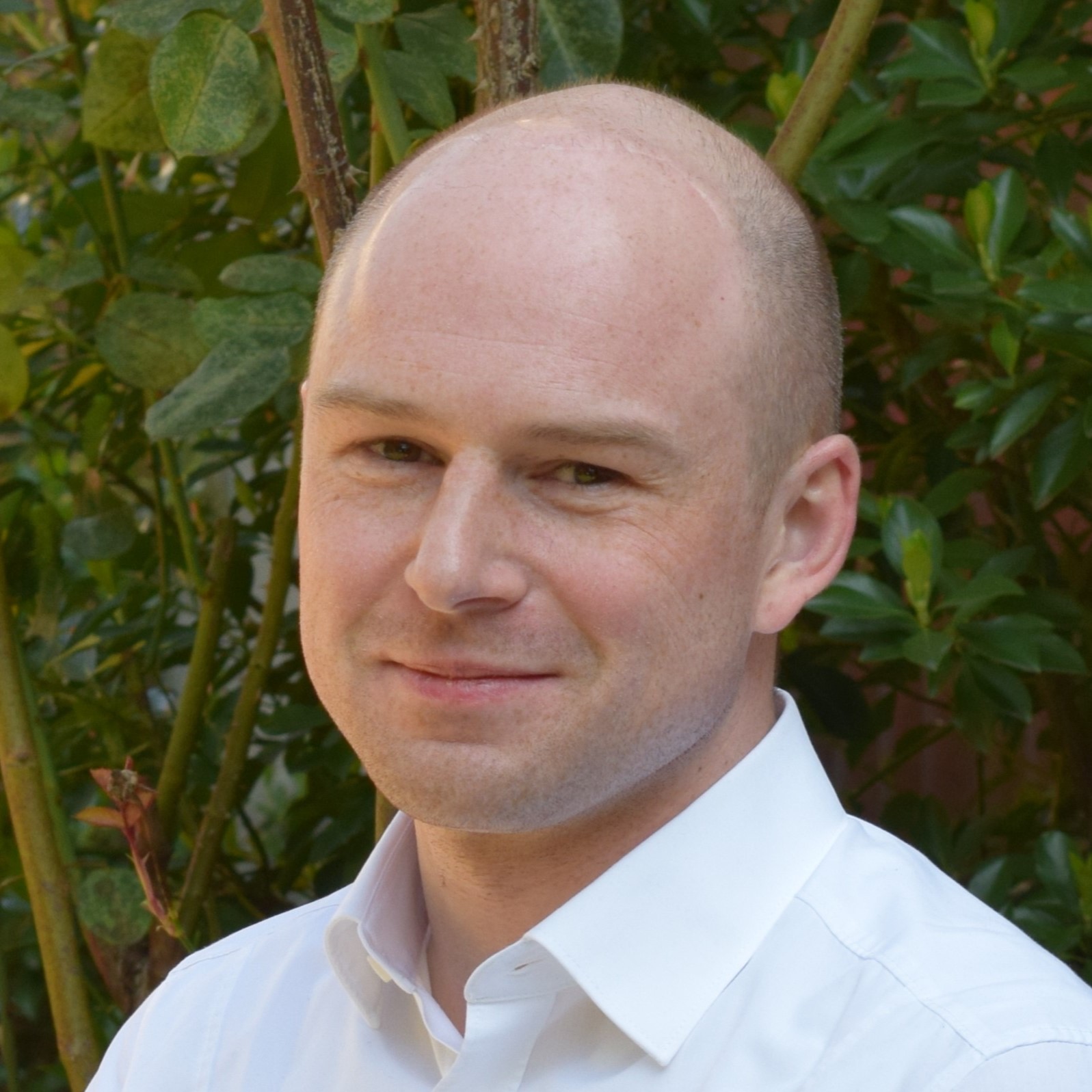 Glatzköpfiger Mann mit weißem Hemd und leichtem Lächeln vor dichtem grünen Blattwerk im Hintergrund.