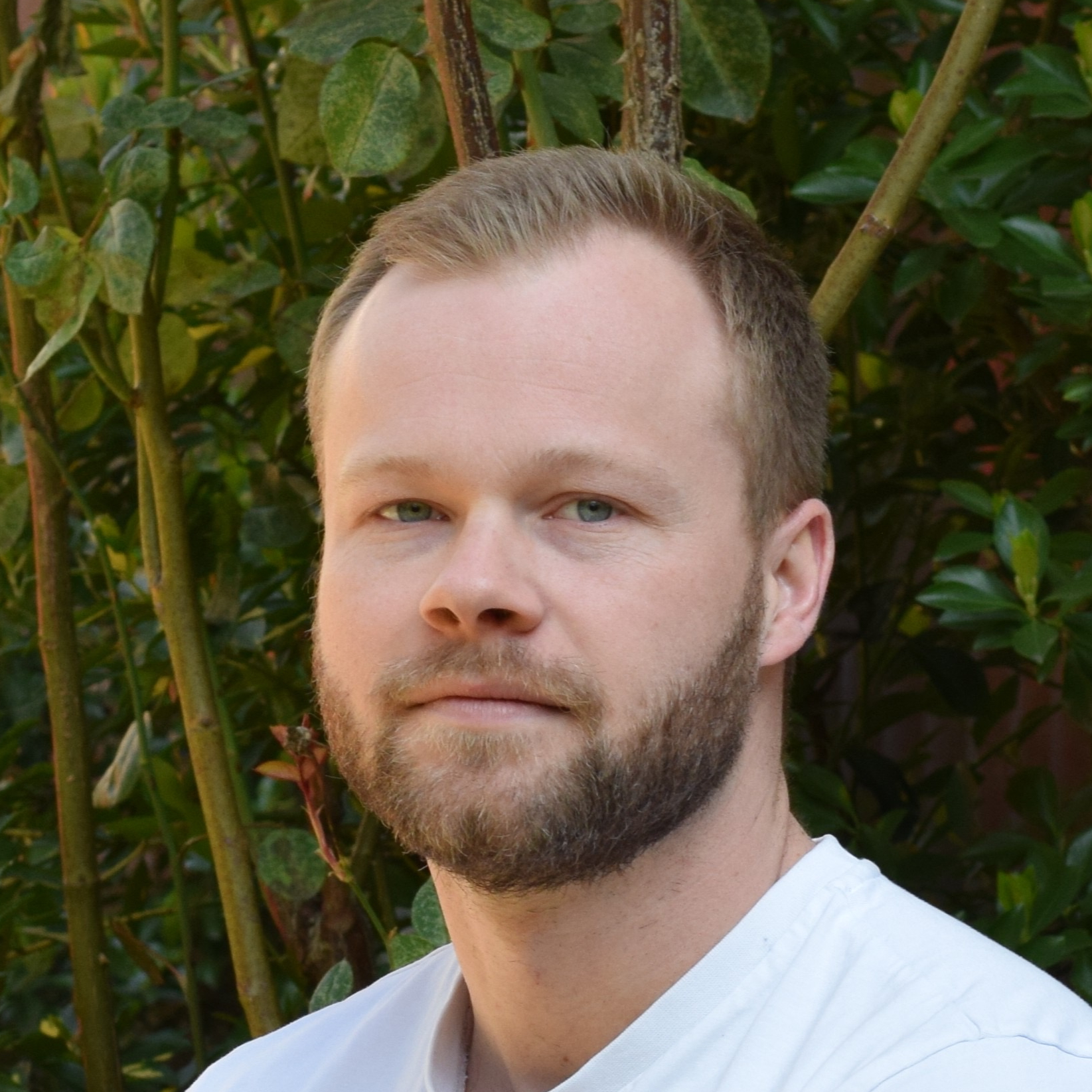 Mann mit braunem Haar und Bart lächelt locker in weißem Shirt vor grünem Hintergrund in die Kamera.