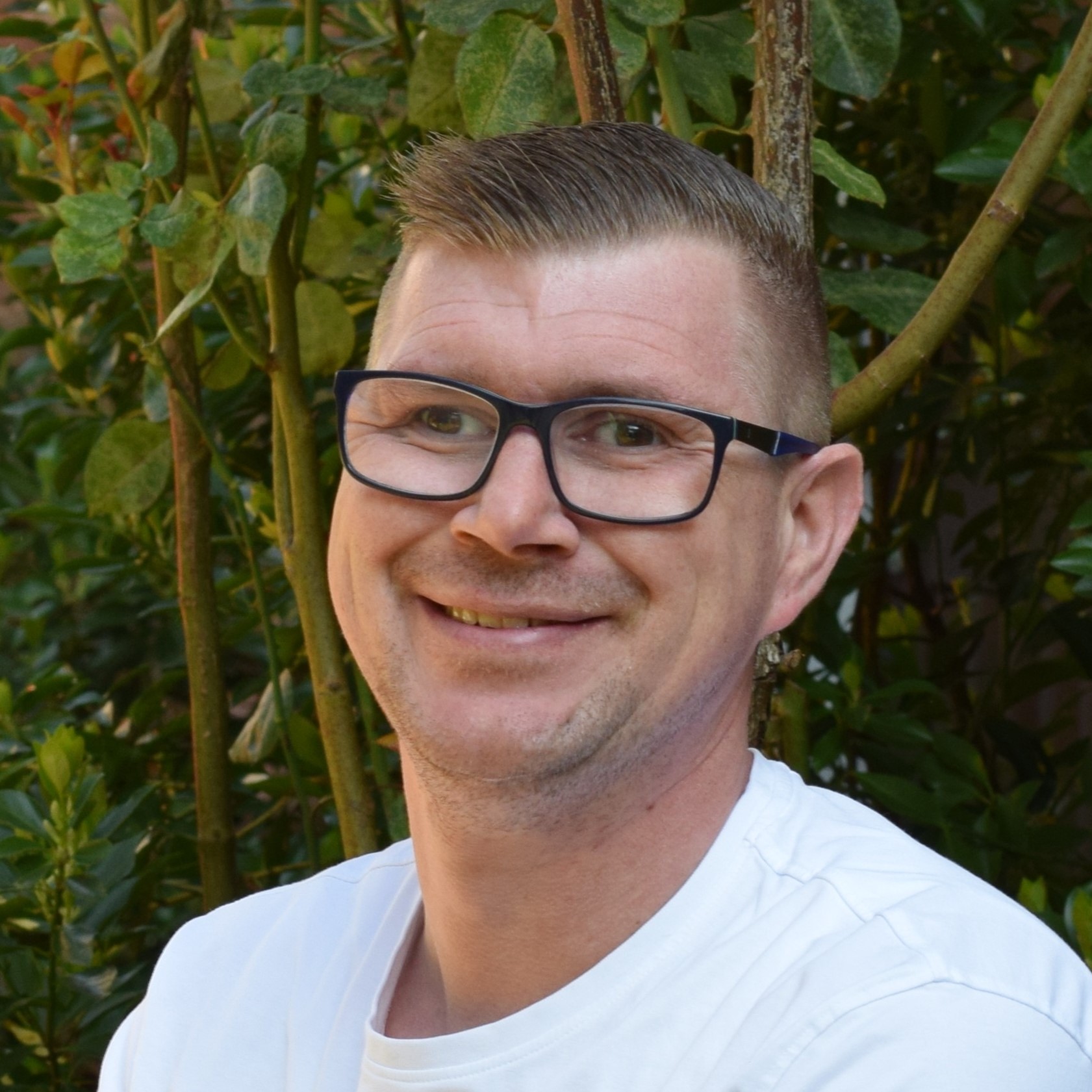 Mann mit kurzem Haar, Brille und weißem Shirt lächelt leicht schräg zur Kamera, Hintergrund mit dichtem Grün.