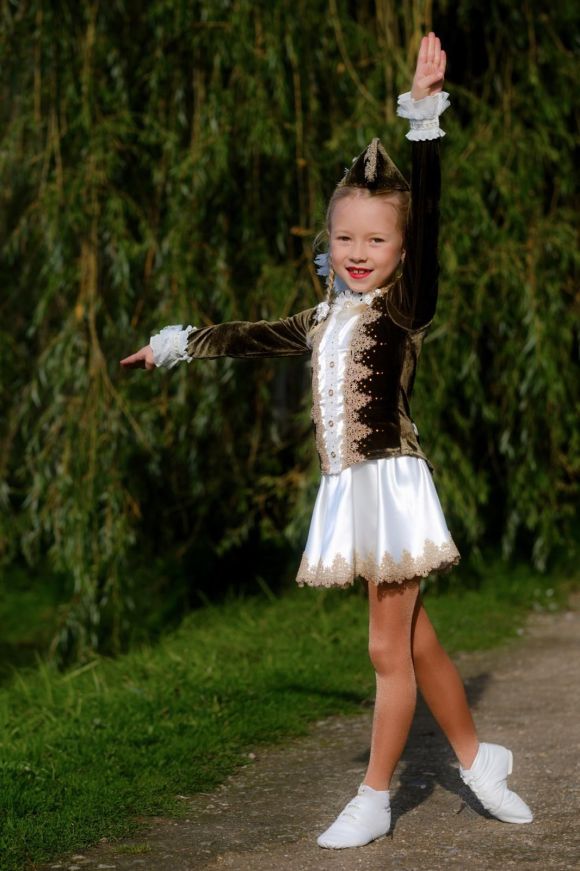 Ein Kind im braun-weißen Tanzkostüm steht mit erhobenem Arm auf einem Weg, hinter ihr ein Weidenbaum.