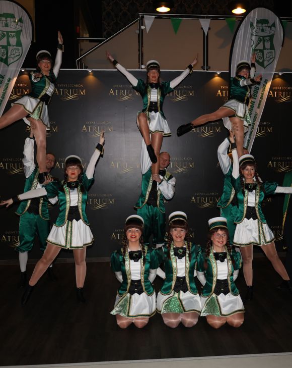 Tanzgruppe in grünen Kostümen bildet akrobatische Pyramiden vor schwarzer Bühne mit „Atrium“-Logo.