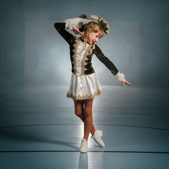 Ein Kind im braun-weißen Tanzkostüm steht mit erhobenem Arm auf einem Weg, hinter ihr ein Weidenbaum.