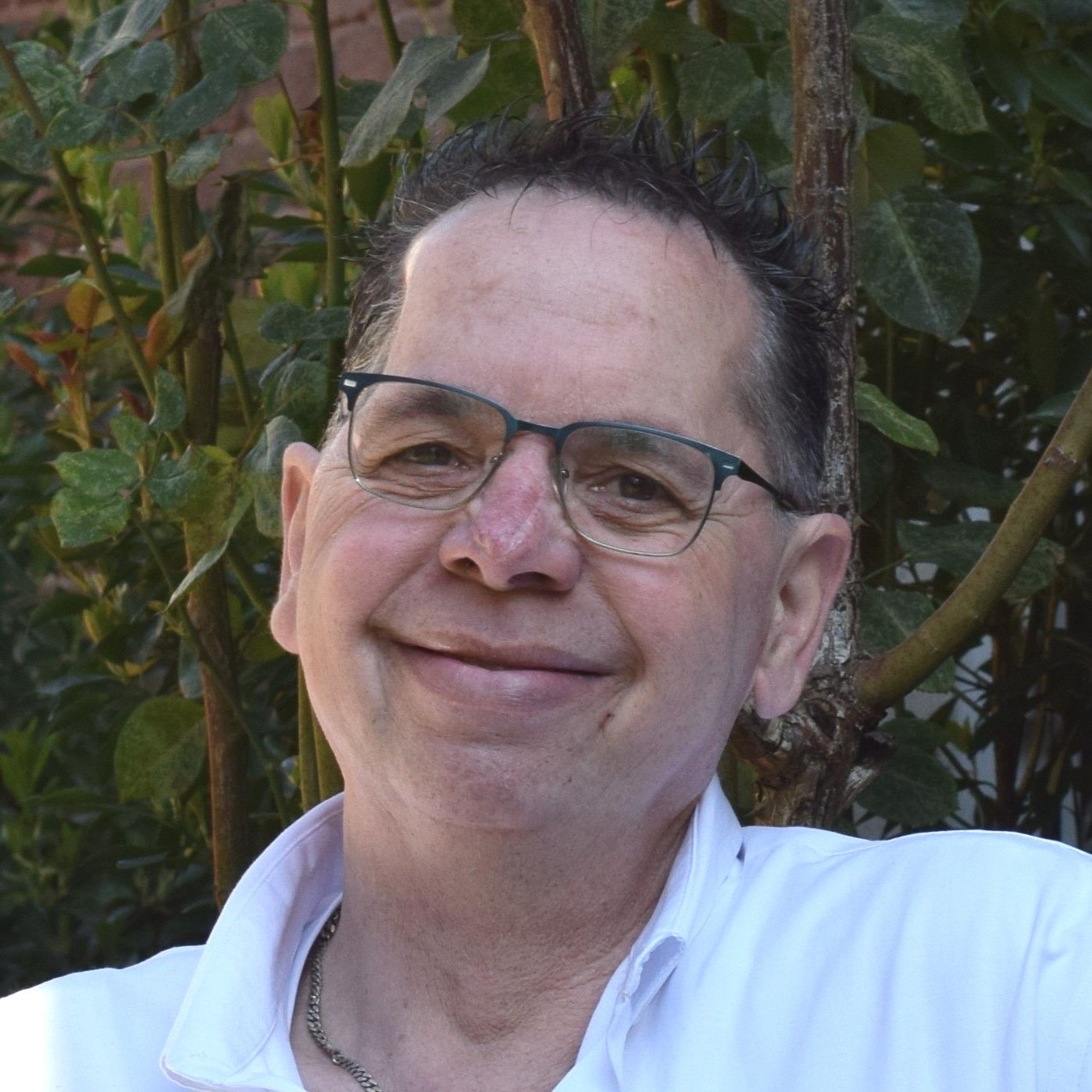 Mann mit Brille und kurzem dunklem Haar lächelt herzlich in weißem Hemd vor grüner Hecke.