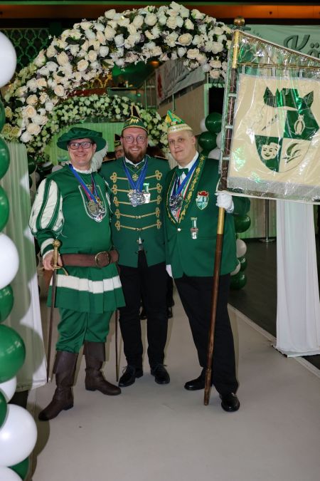 Drei Mitglieder der GOK 1906 e.V. in Karnevalskleidung posieren bei einer Sitzung mit einem Schild ihres Vereins.